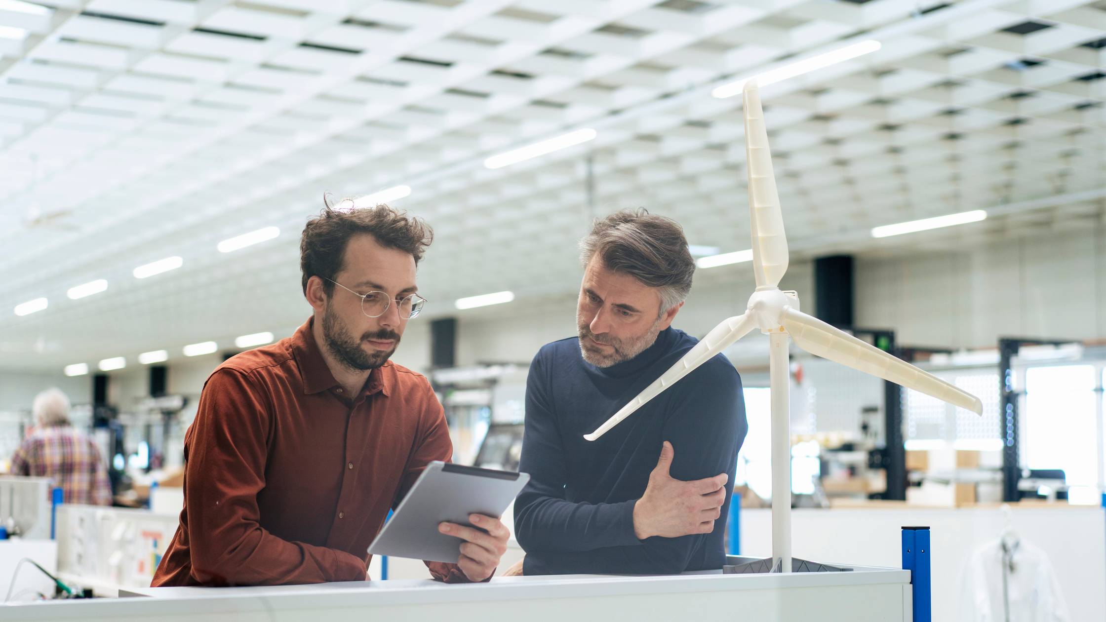 Zwei Männer schauen auf ein digitales Tablet in einem hellen weißen Raum mit einem Turbinenmodell im Vordergrund